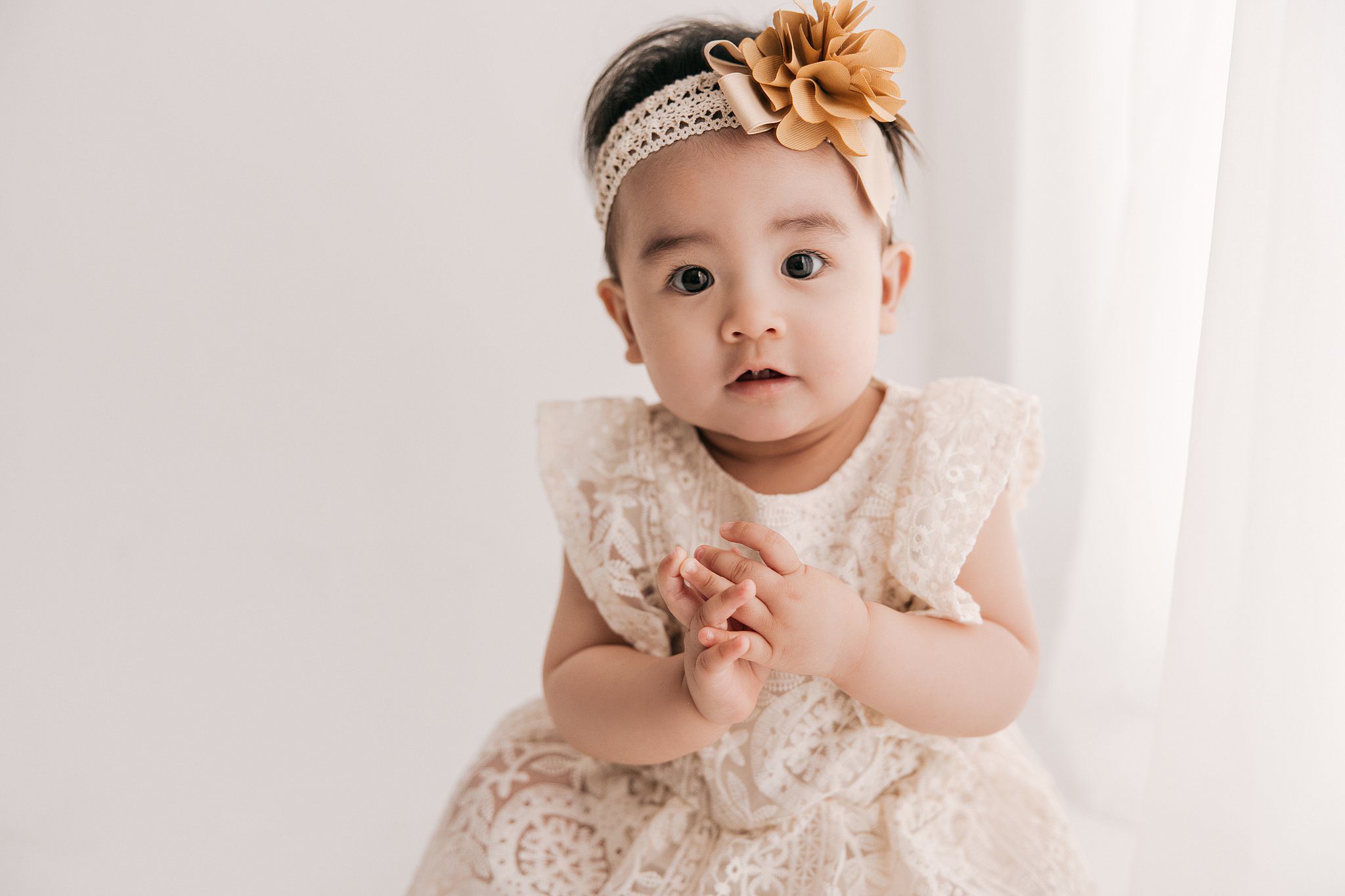 A toddler girl in a cream lace dress sits on the floor of a studio under a window