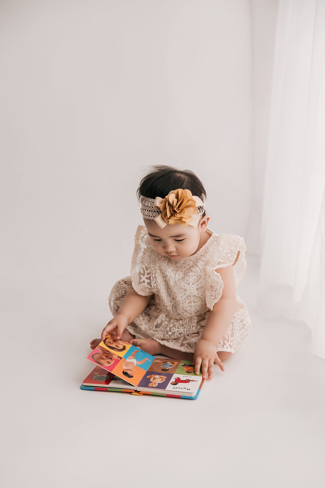 A toddler girl in a lace dress sits on a studio floor turning pages of a picture book after meeting nannies raleigh